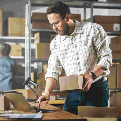 Man with beard holds packaged box in warehouse while working on computer