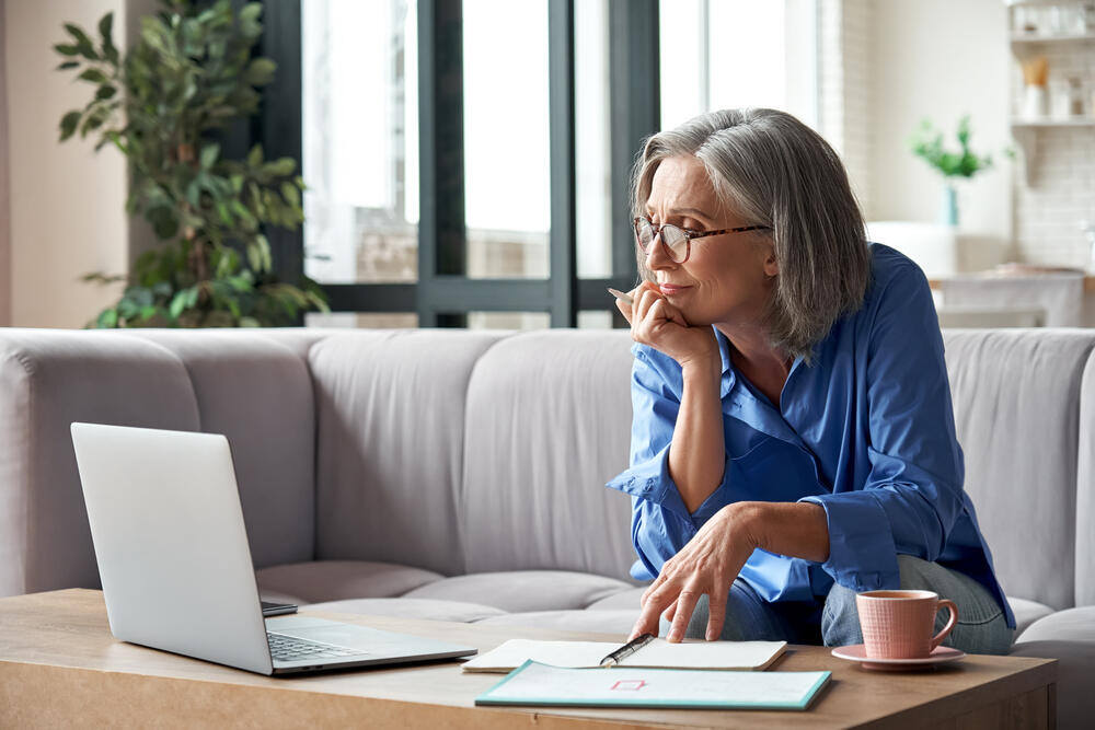 Older woman with grey hair in blue shirts takes notes on notepad while watching webinar on couch