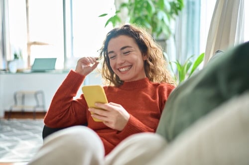 Young woman lounges on couch smiling and checking social media on phone.