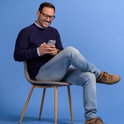 Middle age man sits in chair holding phone and smiling at screen.