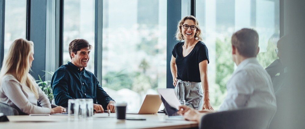 Woman manager stands and smiles at colleagues sitting around conference table in window-lit room.